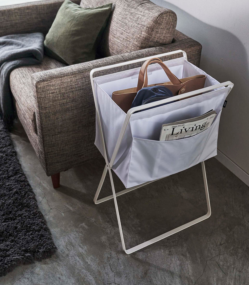 View 20 - A white canvas hamper holding a purse and backpack. Magazines are seen peeking out of a side-pocket. A brown couch with a green throw pillow and gray rug can be seen in the background.