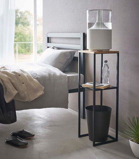 Pedestal Stand by Yamazaki Home in black in a bedroom holding a humidifier and books, and a waste bin underneath. view 11