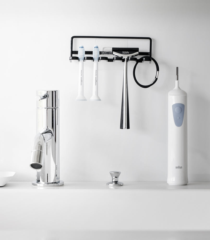 View 9 - Yamazaki Home's black Traceless Adhesive Toothbrush Holder on a bathroom wall, holding two toothbrushes and a razor beside a faucet.
