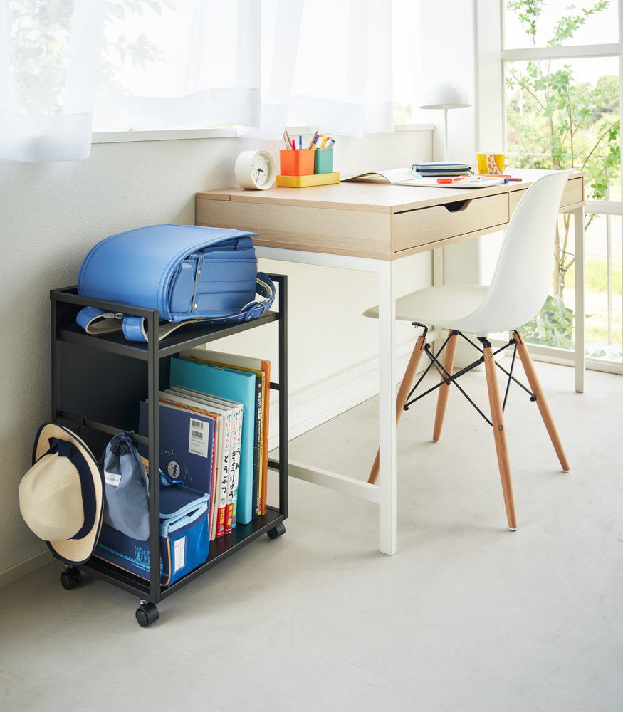 View 13 - Two-level black steel storage cart with casters is shown next to a desk. A backpack is shown on the upper shelf and the lower shelf houses books and a small bag. Three hooks are installed on one side, holding a hat and a small bag. One side of the cart has a wall to hide storage when flipped.