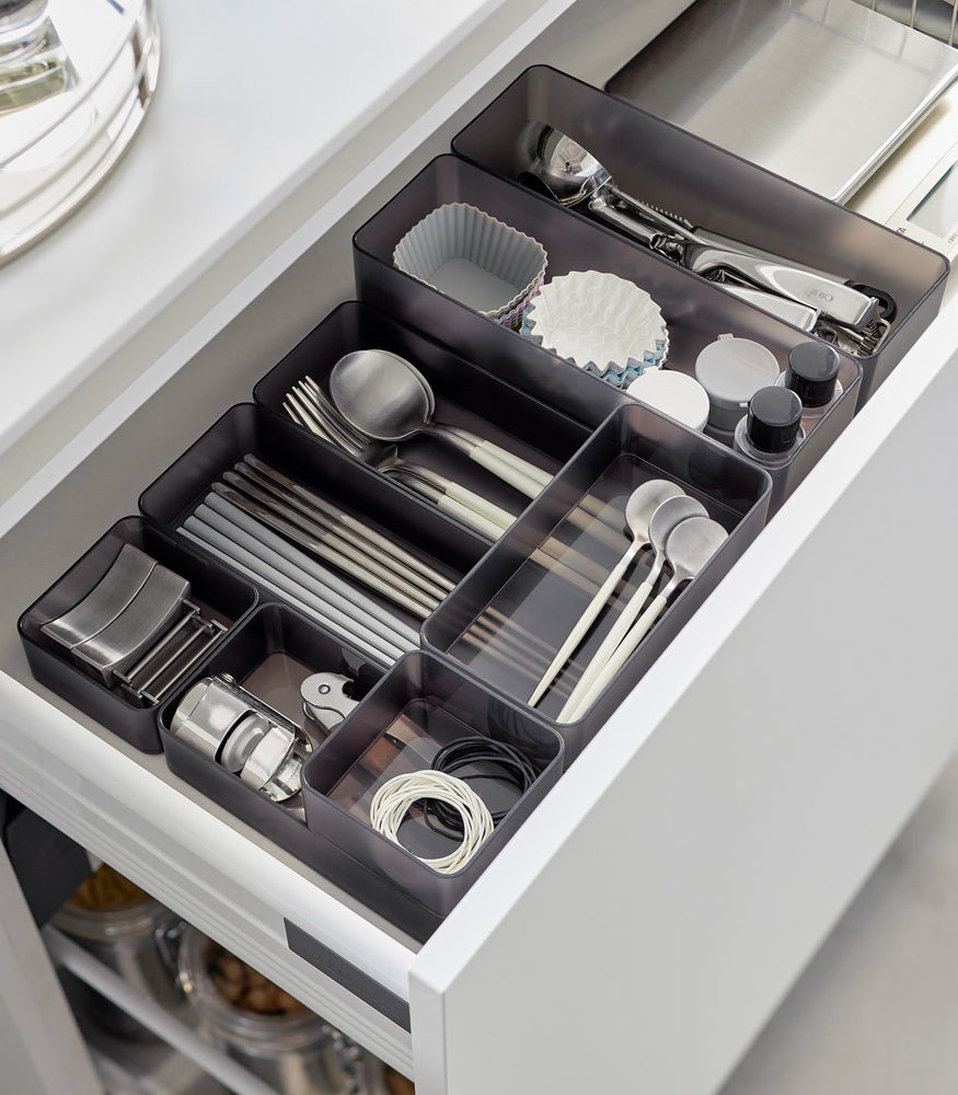 View 44 - Four different sizes of stackable clear organizers in black—small, medium, large, and deep—are neatly placed in a kitchen drawer. They effectively organize various items such as cutlery, bottle openers, and small trays, showcasing their versatility and stackability