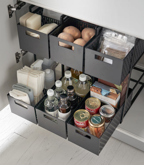 Three two-tier black storage bins are displayed in a lower cabinet. The first houses cleaning supplies, the other two hold pantry items. All levels of the storage bin are pulled out mid-way to show its storage possibilities.