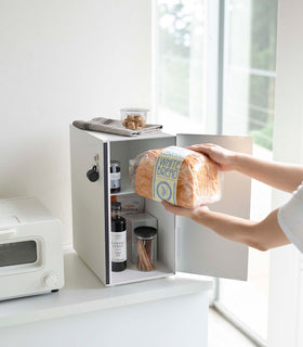 An angled view of a person’s arms putting bread into a white metal vertical breadbox placed on top of a white kitchen counter. The door is swung open from the right and a magnetic strip is seen opposite of its position. Coffee Syrup, cinnamon sticks in a clear container, and other pantry items are seen in the open breadbox. A magnetic hook is attached to its side. On top is a folded tea towel and clear package of treats. Slightly off frame is a white microwave oven. view 19
