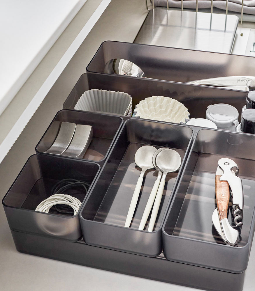 View 61 - Close-up image of four different sizes of stackable clear organizers in black—small, medium, large, and deep—neatly placed in a kitchen drawer. They effectively organize various items such as cutlery, bottle openers, and small trays, showcasing their versatility and stackability.