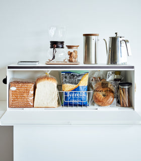 Frontal view of white Yamazaki Home Bread Box - Wide open with bread and snacks inside and coffee equipment on top view 5