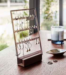 An acrylic translucent mauve earring holder with a rectangular wooden base are displayed on a dark wood dresser. The acrylic holder has upward pointed hooks and slots placed in an interchangeable pattern. Hanging from the hooks are chained necklaces, and in the slots are various earrings. On the surface of the dresser, in front of the earring holder, are two rings next to a decorative catch-all plate. view 9