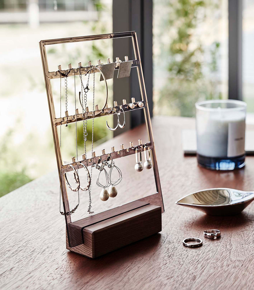 View 9 - An acrylic translucent mauve earring holder with a rectangular wooden base are displayed on a dark wood dresser. The acrylic holder has upward pointed hooks and slots placed in an interchangeable pattern. Hanging from the hooks are chained necklaces, and in the slots are various earrings. On the surface of the dresser, in front of the earring holder, are two rings next to a decorative catch-all plate.