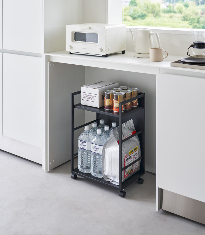 View 11 - Two-level black steel storage cart with casters is shown in an opening of a recessed cabinet. Cart houses a box and six cans of food on the upper shelf, the lower shelf holds a large bag of food with 6 liters of bottled water. One side of the cart has a wall to hide storage.