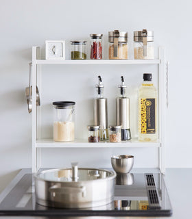 Front view of Two-Tier Countertop Rack by Yamazaki Home in white by a gas range storing various seasonings. view 4