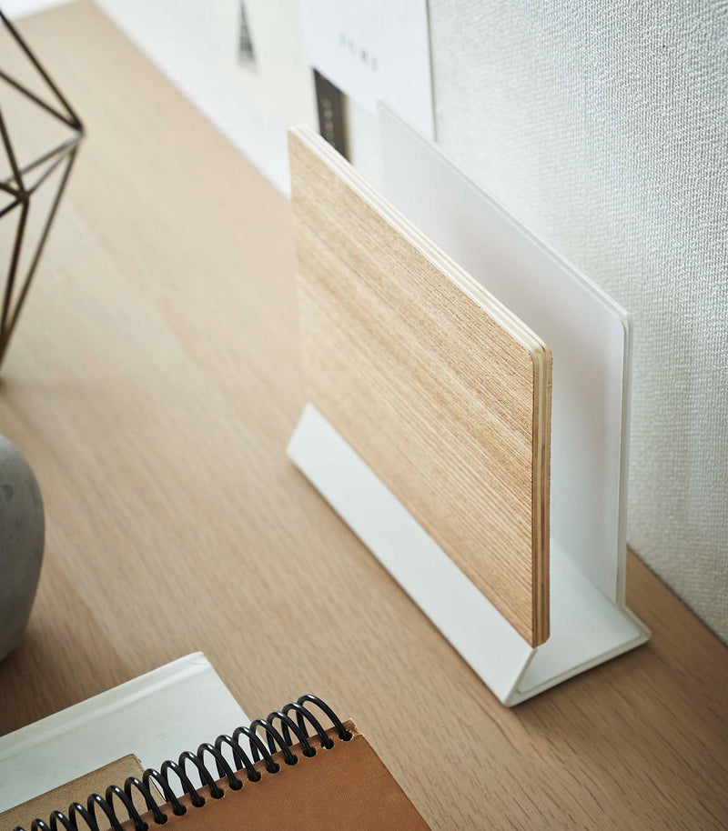 A side-view profile of a narrow wooden stand with a white metal base positioned by books on top of a desk.