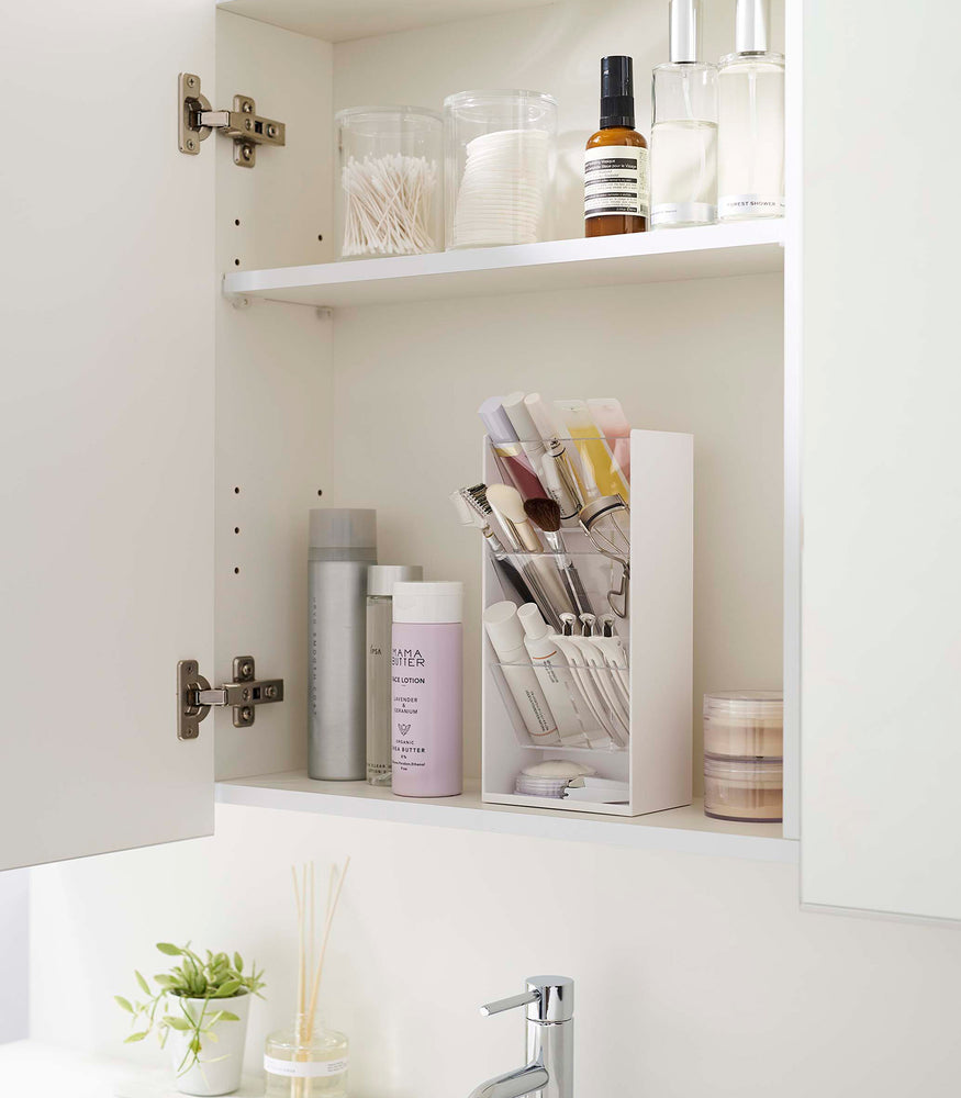 View 4 - An angled view of an opened white medicine cabinet. Sunlight is focused on the right upper corner. The top half of the cabinet holds face serums and two clear containers with cotton swabs and rounds.