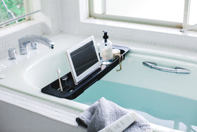 Side view of black Expandable Bathtub Caddy holding cleaning items and tablet in bathroom by Yamazaki Home. view 8