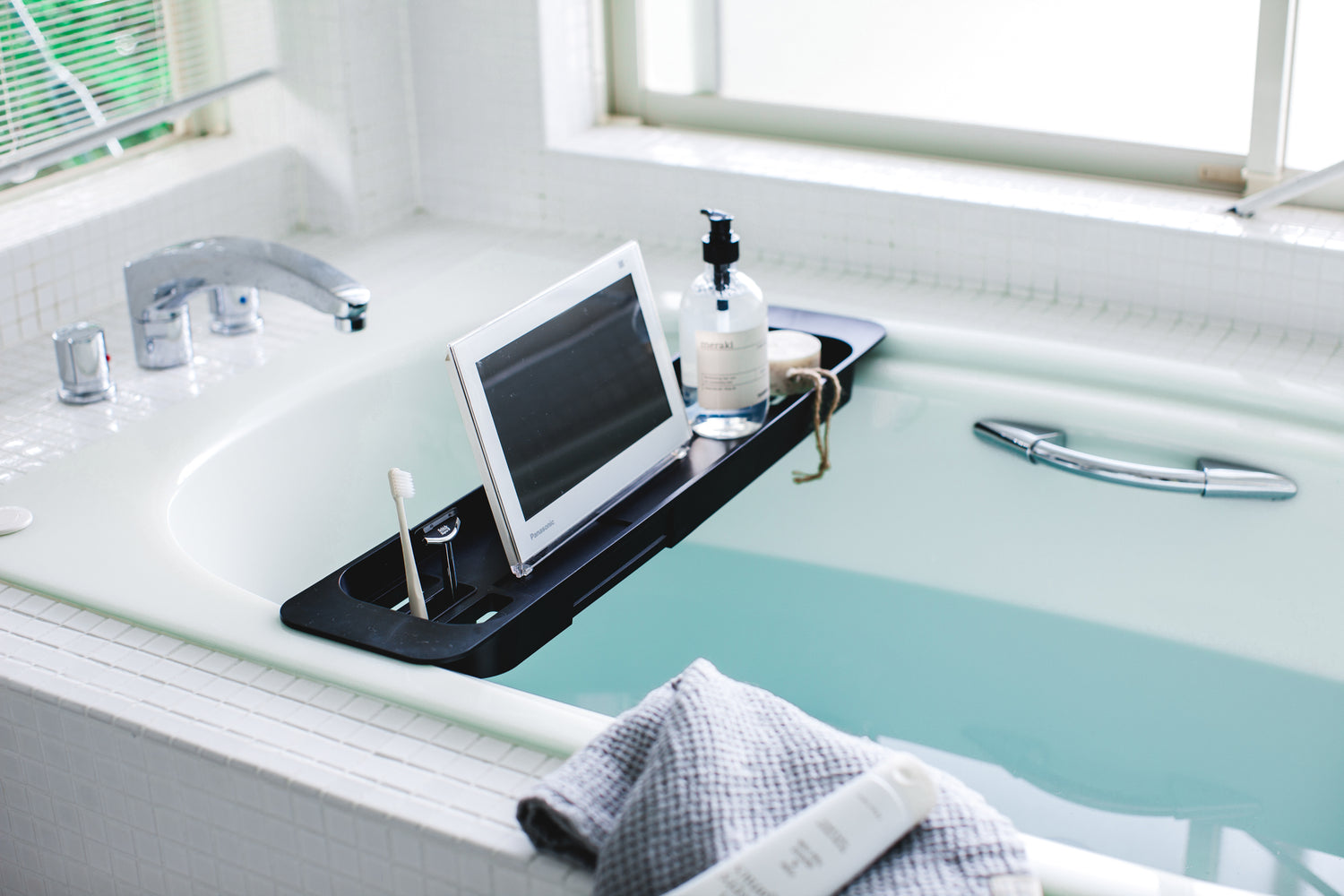 View 8 - Side view of black Expandable Bathtub Caddy holding cleaning items and tablet in bathroom by Yamazaki Home.