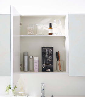 A white medicine cabinet is open to display the inside contents. Sunlight is focused on the right upper corner. Below is a bathroom sink with a silver faucet. On the sinks ledge is a small plant and oil diffuser. view 15