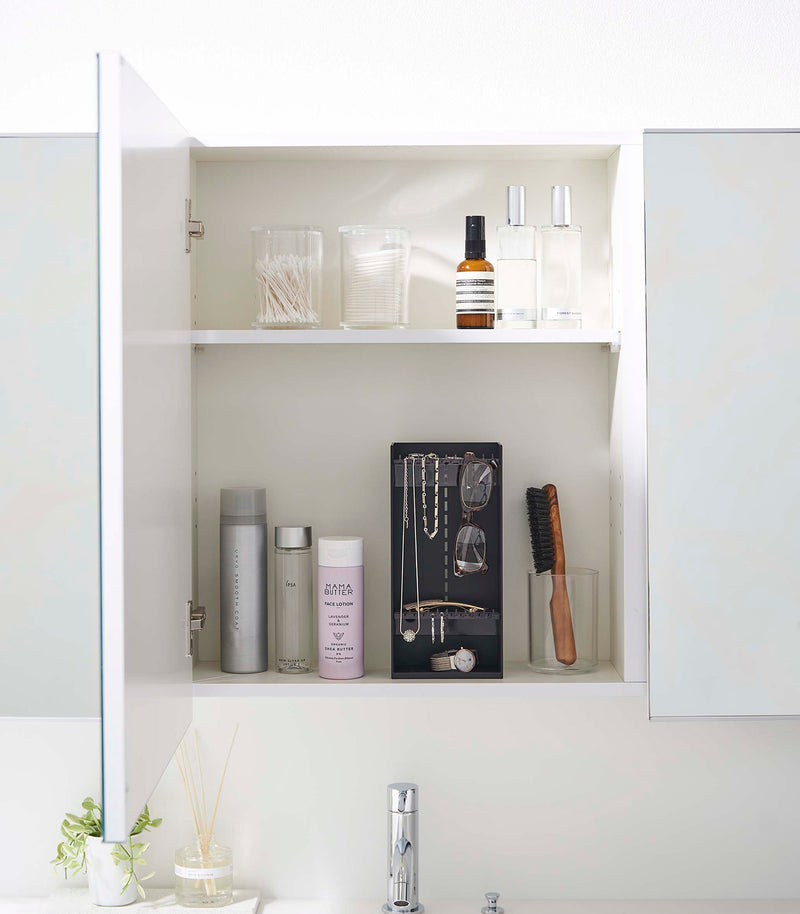 A white medicine cabinet is open to display the inside contents. Sunlight is focused on the right upper corner. Below is a bathroom sink with a silver faucet. On the sinks ledge is a small plant and oil diffuser.
