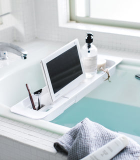 White Expandable Bathtub Caddy holding tablet and cleaning products in bathroom by Yamazaki Home. view 2