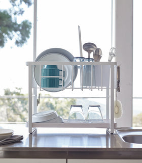 Front view of white Two-Tier Dish Rack holding plates, cups, and silverwear on kitchen counter by Yamazaki Home. view 6