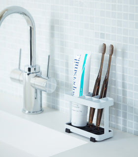 White Toothbrush Stand displaying toothpaste and toothbrushes on bathroom sink counter by Yamazaki Home. view 3