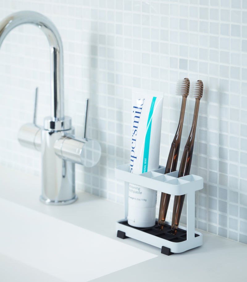 White Toothbrush Stand displaying toothpaste and toothbrushes on bathroom sink counter by Yamazaki Home.