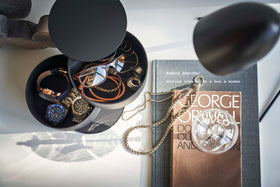 A bird’s-eye view of a black two-tier swivel accessory holder on a white dresser. To the right of it are a stack of books and two strewn gold chains.  The accessory holder’s tiers and lid are swiveled opened so the inside contents can be seen. In the first tier are a pair of reading glasses, two silver statement rings and tan corded bracelets. The second tier holds three watches of different styles. view 15