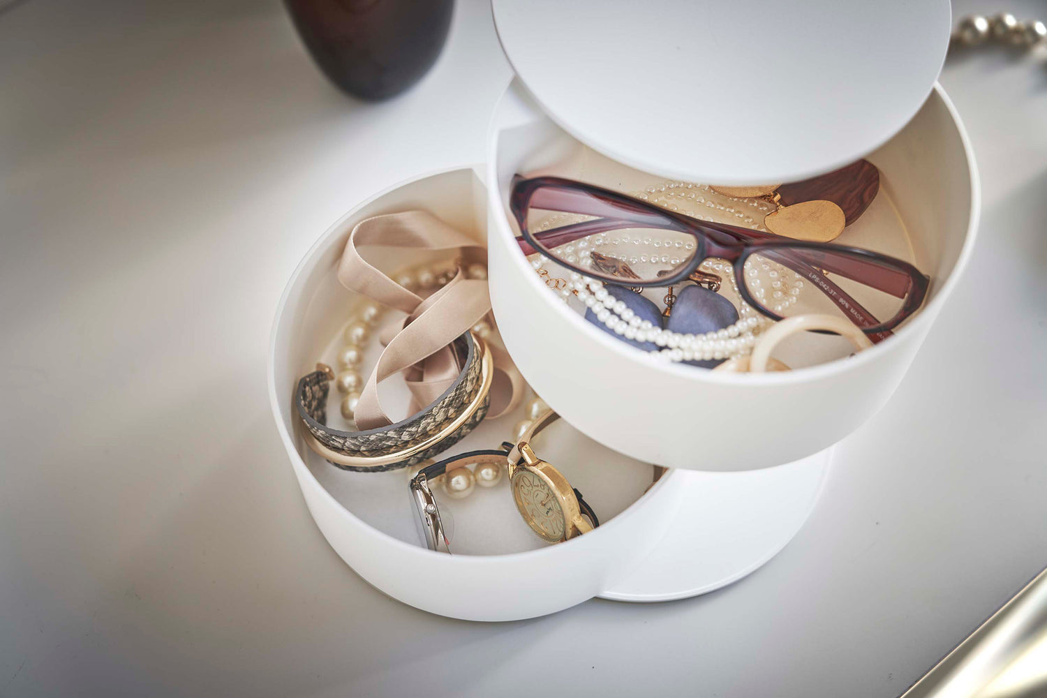 View 6 - A detailed view of a two-tier white swivel accessory holder. The tiers and lid are swiveled open so the inside contents can be seen. In the first tier are a pair of reading glasses, earrings, and other various jewelry. The second tier holds a watch and an assortment of bracelets.