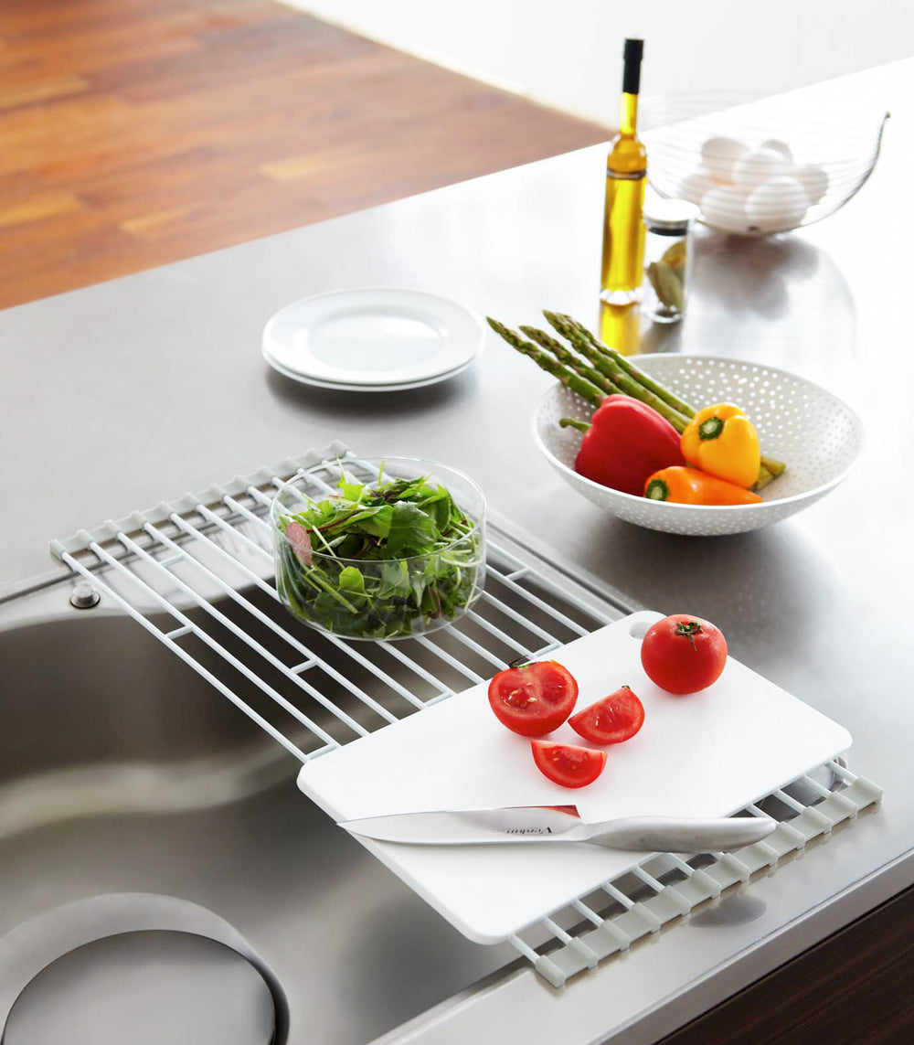 Over-the Dish Drainer holding cutting board and vegetables on sink counter by Yamazaki Home.