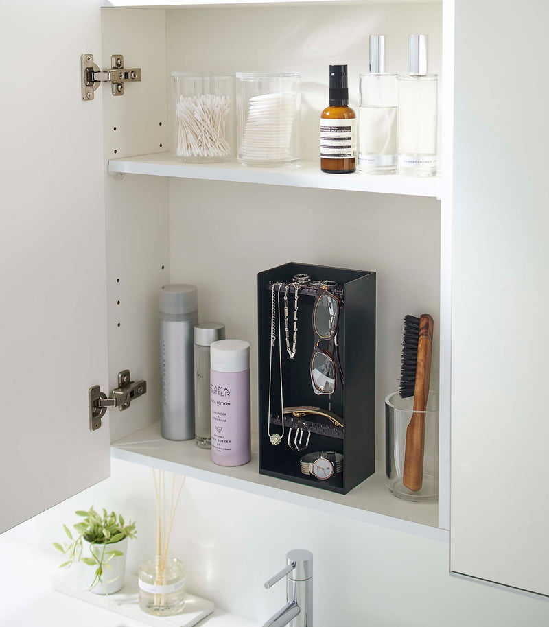 A white medicine cabinet is open to display the inside contents. Sunlight is focused on the right upper corner. Below is a bathroom sink with a silver faucet. On the sinks ledge is a small plant and oil diffuser.