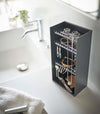 Looking down on top of a white bathroom counter is a black resin rectangular jewelry holder with an open face and top with three removable transparent shelves with upward facing hooks along the edge.