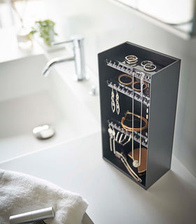 Looking down on top of a white bathroom counter is a black resin rectangular jewelry holder with an open face and top with three removable transparent shelves with upward facing hooks along the edge. view 11