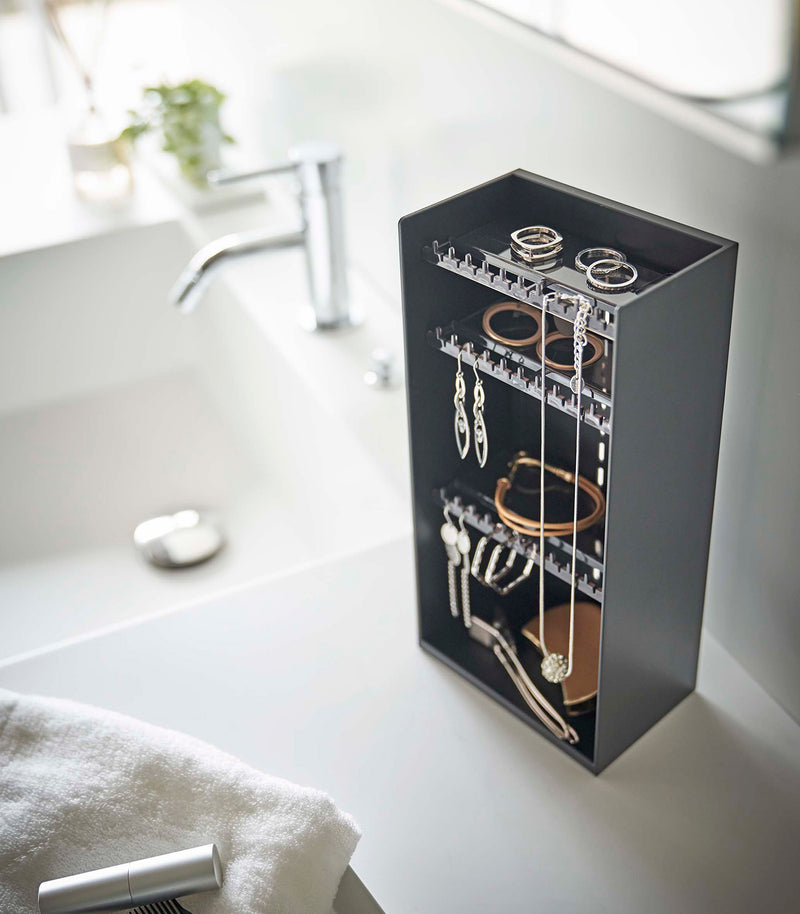 Looking down on top of a white bathroom counter is a black resin rectangular jewelry holder with an open face and top with three removable transparent shelves with upward facing hooks along the edge.