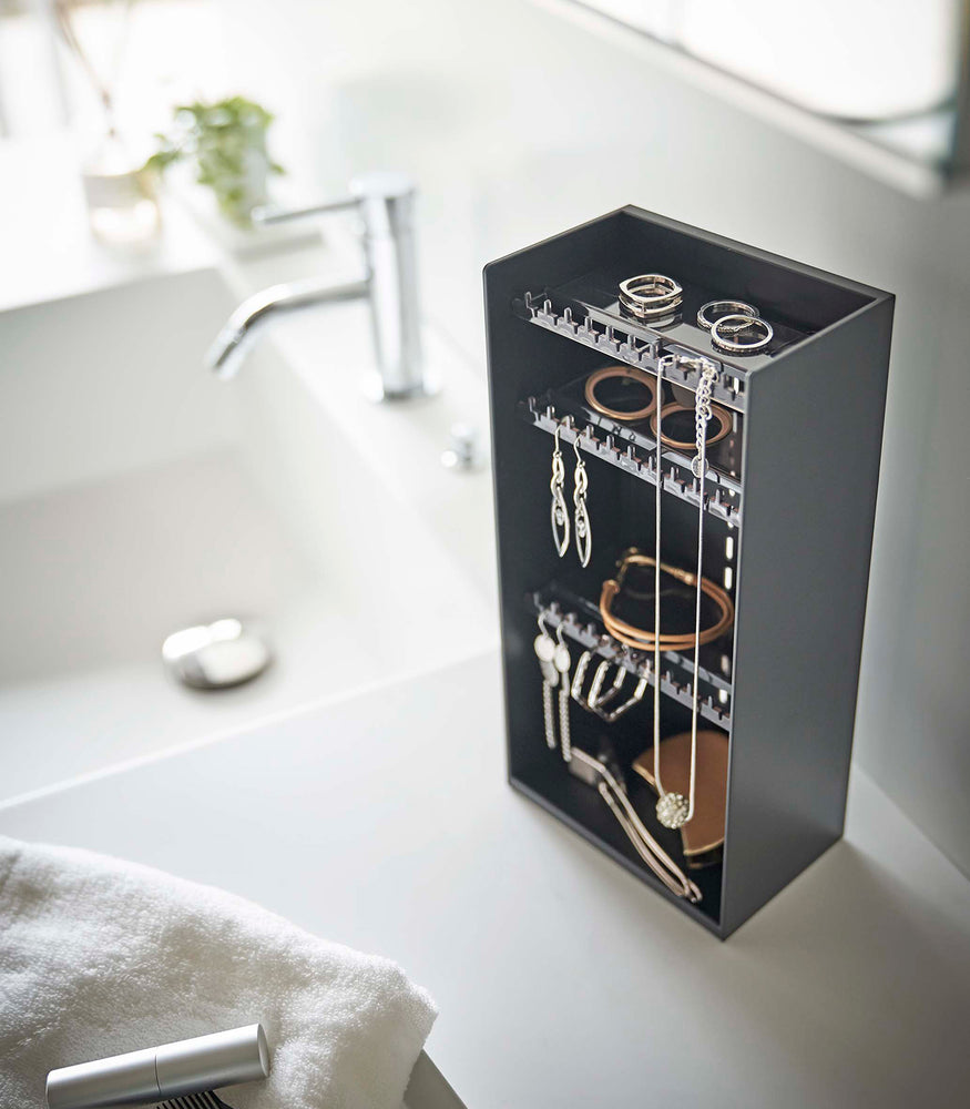 View 11 - Looking down on top of a white bathroom counter is a black resin rectangular jewelry holder with an open face and top with three removable transparent shelves with upward facing hooks along the edge.