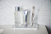 Front view of white Slotted Tray holding toothbrush, toothpaste, and mouthwash on bathroom sink by Yamazaki Home.