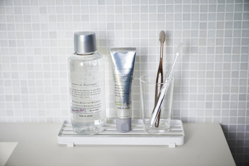 Front view of white Slotted Tray holding toothbrush, toothpaste, and mouthwash on bathroom sink by Yamazaki Home.