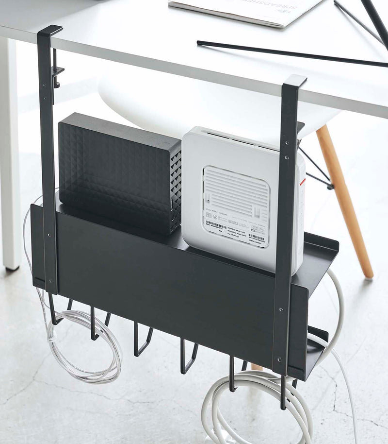 Back view of black Wall-Mount Cable and Router Storage Rack hanging from desk by Yamazaki Home.