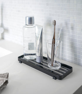 Black Slotted Tray holding toothbrush and beauty items on bathroom sink counter by Yamazaki Home. view 7