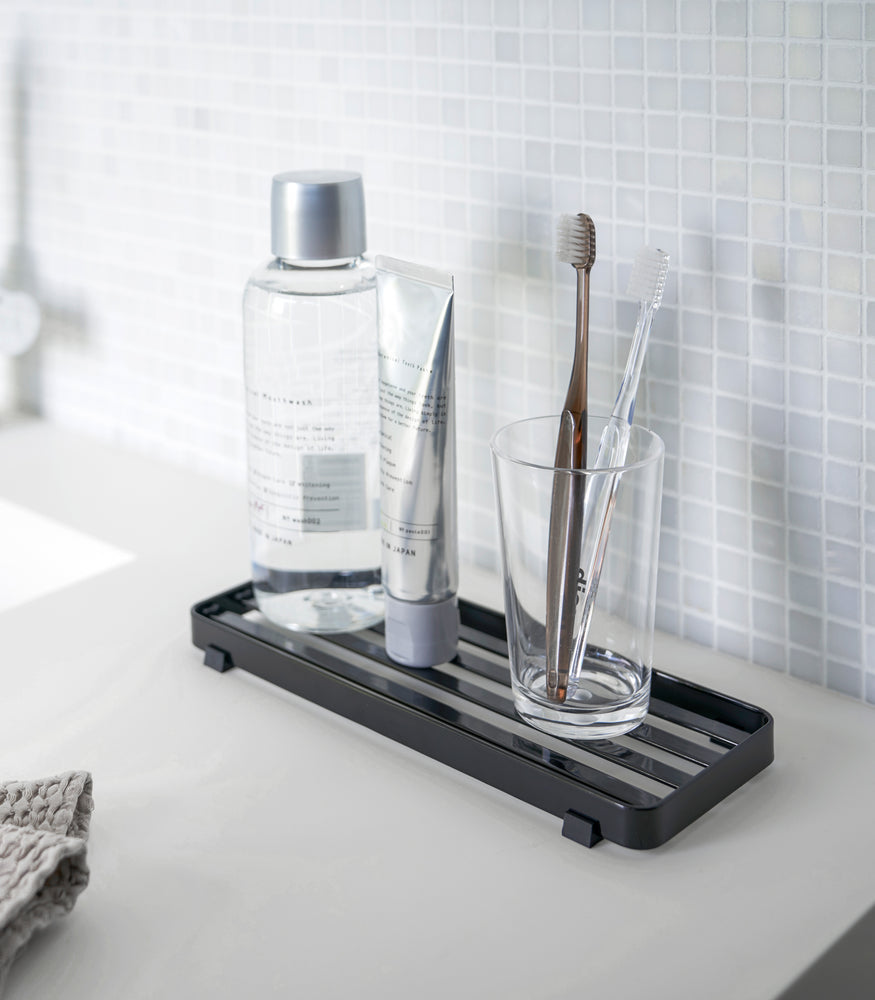 View 7 - Black Slotted Tray holding toothbrush and beauty items on bathroom sink counter by Yamazaki Home.