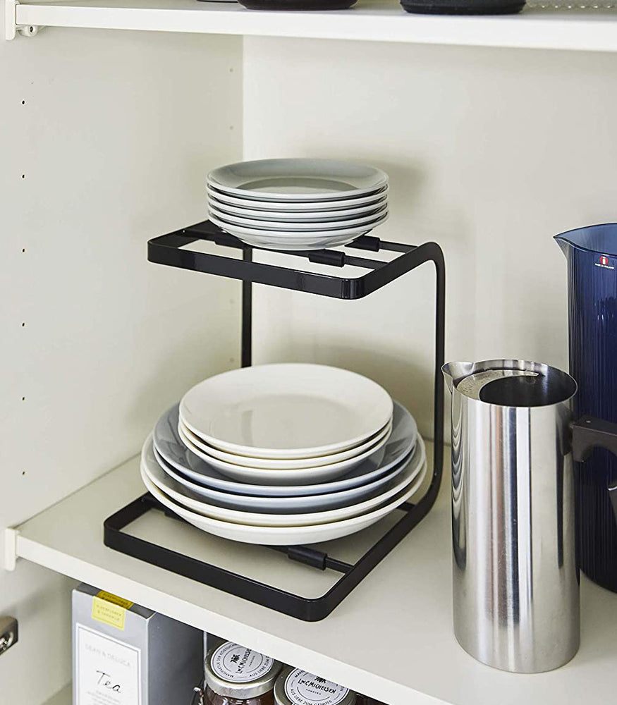 View 11 - Black 2-Tier Pot Holder with Hooks holding plates in kitchen cabinet by Yamazaki Home.