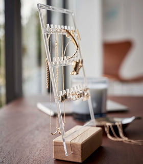 A profile view of a clear acrylic earring holder with a light-colored rectangular wooden base on a dark wood dresser. The acrylic holder has upward pointed hooks and slots placed in an interchangeable pattern. Hanging from the hooks are chained necklaces, and in the slots are various earrings. Out-of-focus behind the product are a leaf-shaped decorative catch-all plate with necklaces strewn inside. view 5
