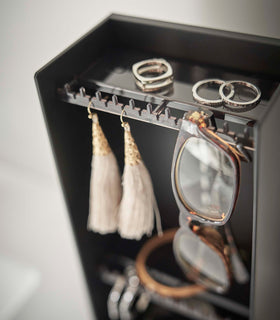 A close-up shot of the corner of a black plastic jewelry organizer. A transparent shelf with an upward facing lip is shown with hooks along the edge. Out-of-focus are three silver rings sitting on the shelf, and hanging on the hooks are a pair of out-of-focus earrings, in focus are the corner of a pair of reading glasses threaded along the hooks edge. view 14