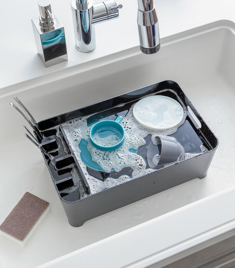 Black Dish Rack in kitchen sink holding water and dishes by Yamazaki Home.
