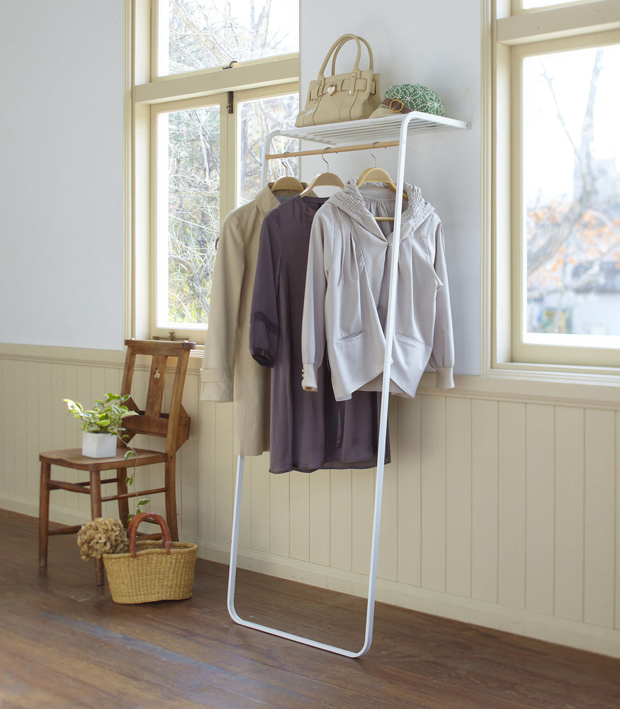 View 2 - White Leaning Coat Rack with Shelf holding purse, hat, and jackets by Yamazaki Home.