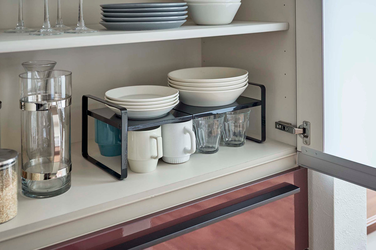 View 11 - Black Expandable Countertop Organizer expanded holding plates above cups in kitchen cabinet by Yamazaki Home.