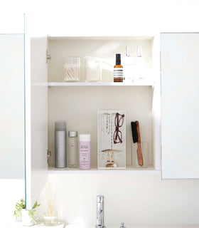 A white medicine cabinet is open to display the inside contents. Sunlight is focused on the right upper corner. Below is a bathroom sink with a silver faucet. On the sinks ledge is a small plant and oil diffuser. A white shelf divides the cabinet. view 6