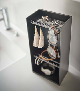 Looking down on top of a white bathroom counter is a black resin rectangular jewelry holder with an open face and top with two removable transparent shelves with upward facing hooks along the edge. view 13