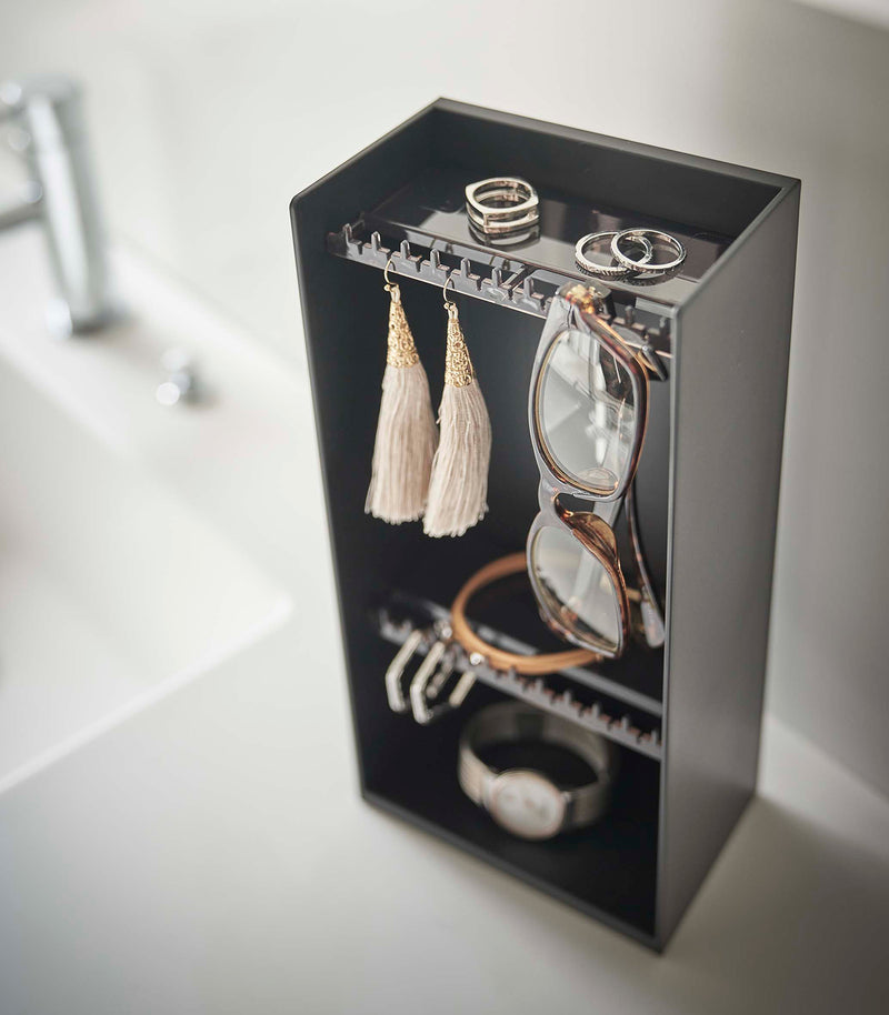 Looking down on top of a white bathroom counter is a black resin rectangular jewelry holder with an open face and top with two removable transparent shelves with upward facing hooks along the edge.
