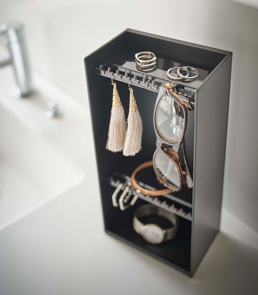 View 13 - Looking down on top of a white bathroom counter is a black resin rectangular jewelry holder with an open face and top with two removable transparent shelves with upward facing hooks along the edge.