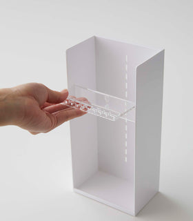 On a white surface is a white resin rectangular jewelry holder with an open face and top with a removable transparent shelf with upward facing along the edge. The bottom of the organizer has a small upward facing lip. A male hand pulls a transparent tray to adjust the location within the jewelry organizer. view 8