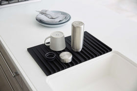Black Folding Dish Drainer Mat holding cup and bottle on sink counter by Yamazaki Home. view 10