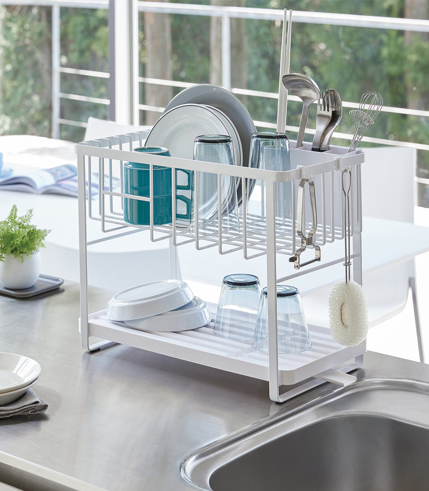 View 3 - White Two-Tier Dish Rack holding plates, cups, and silverwear next to kitchen sink by Yamazaki Home.