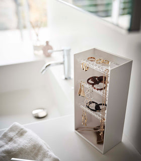 Looking down on top of a white bathroom counter is a white resin rectangular jewelry holder with an open face and top with three removable transparent shelves with upward facing along the edge. view 2